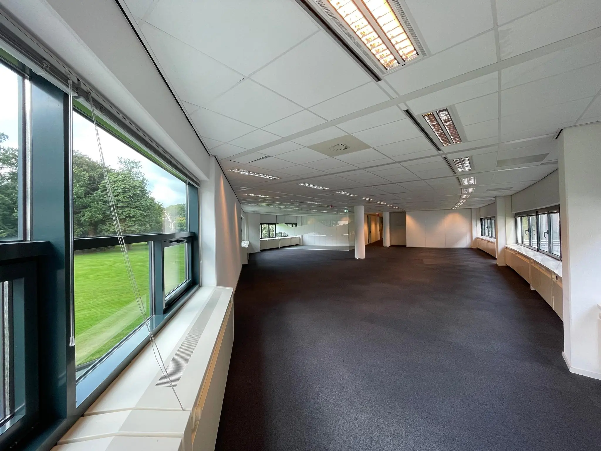 Lege kantoorruimte in kantoorgebouw Sparrenheuvel met grote ramen en uitzicht op een groen grasveld.