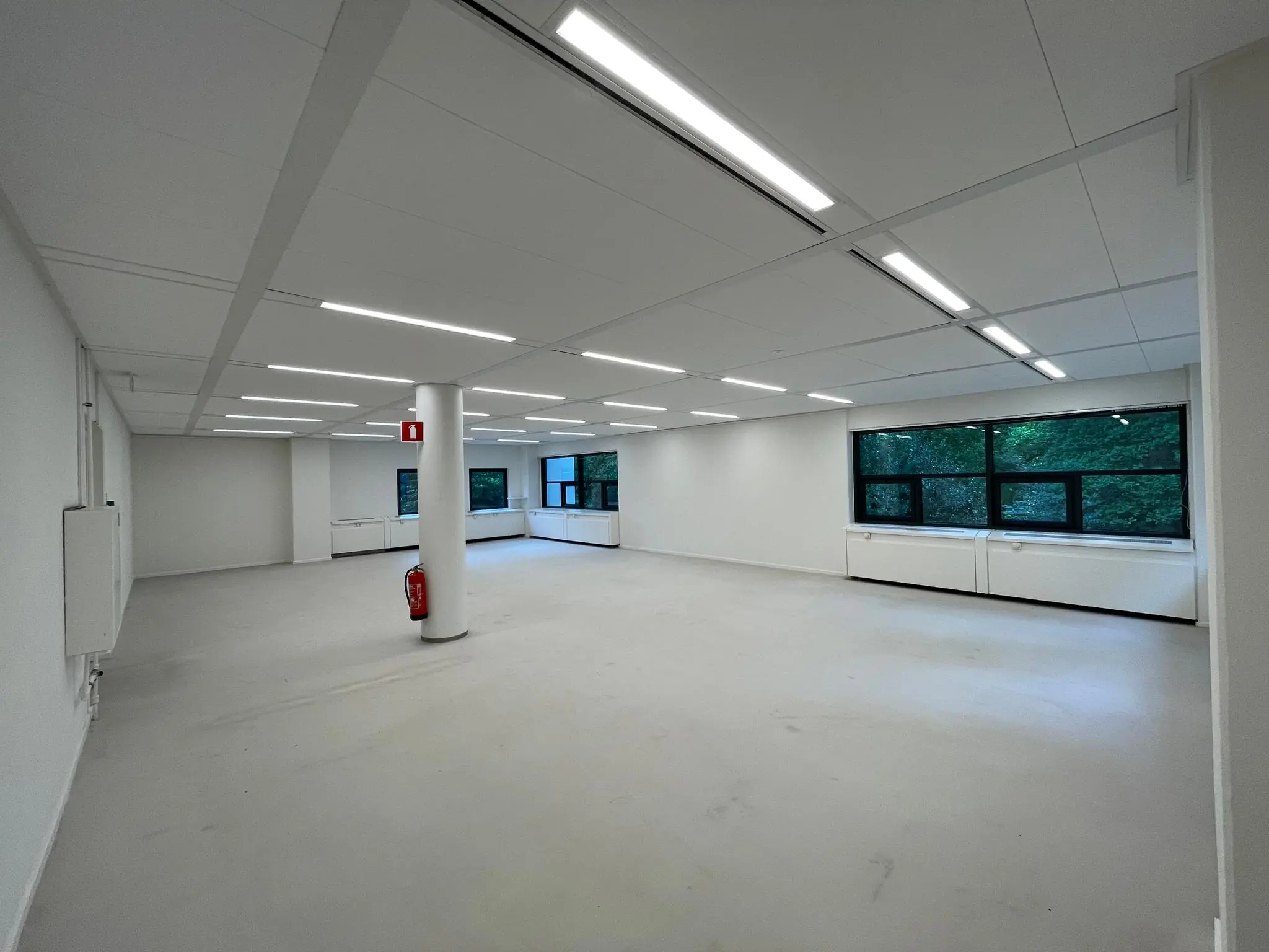 Leeg kantoorruimte in gebouw Sparrenheuvel met witte muren, systeemplafond en grote ramen met uitzicht op groen.
