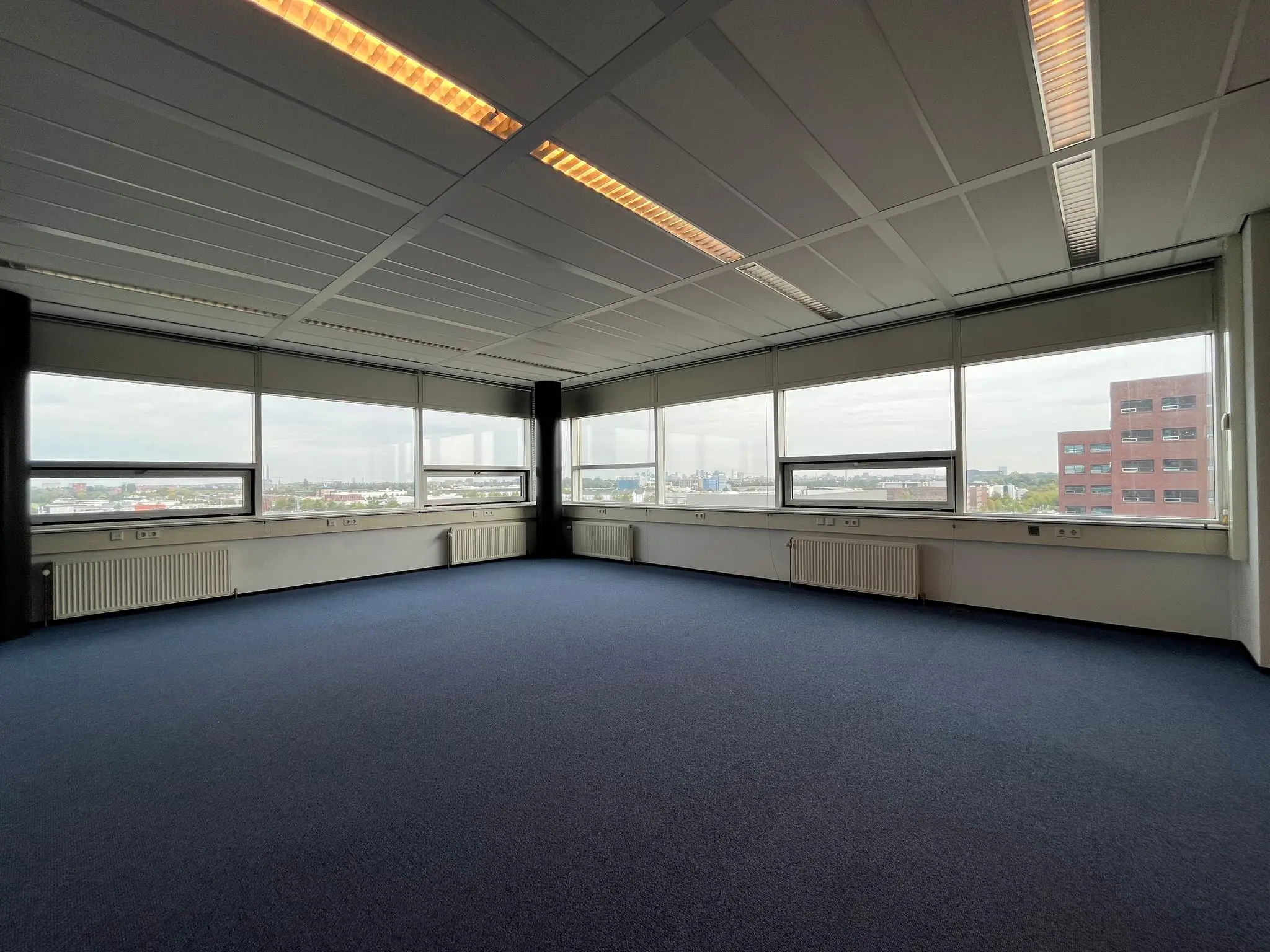 Licht en ruim leeg kantoor met blauwe vloerbedekking en uitzicht op bedrijventerrein Rijnzathe.
