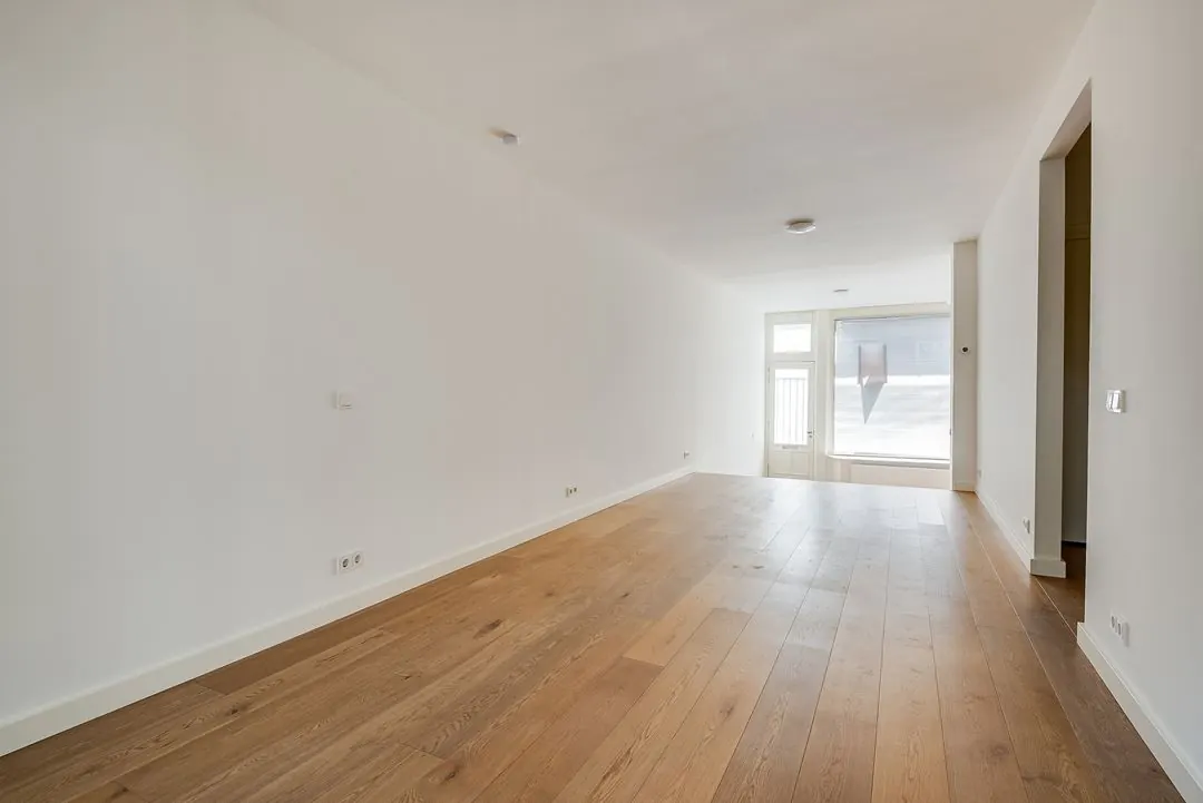 Lichte, lege woonkamer met houten vloer aan de Domselaerstraat, met zicht op een raam en deur aan de voorzijde.