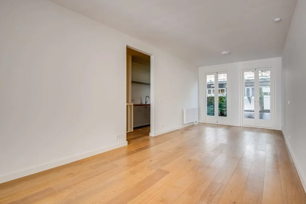 Lichte woonkamer in een woning aan de Domselaerstraat met houten vloer, open doorgang naar de keuken en openslaande deuren naar de tuin.