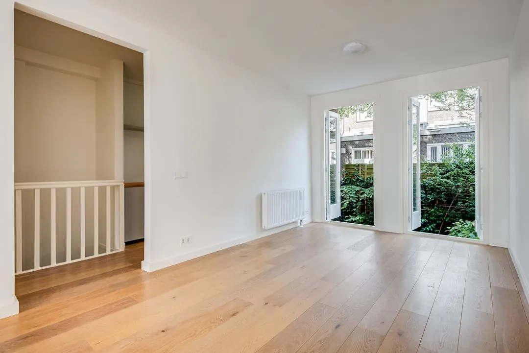 Lichte, lege kamer met houten vloer, openslaande deuren naar tuin en wit geschilderde muren in een woning aan de Domselaerstraat.