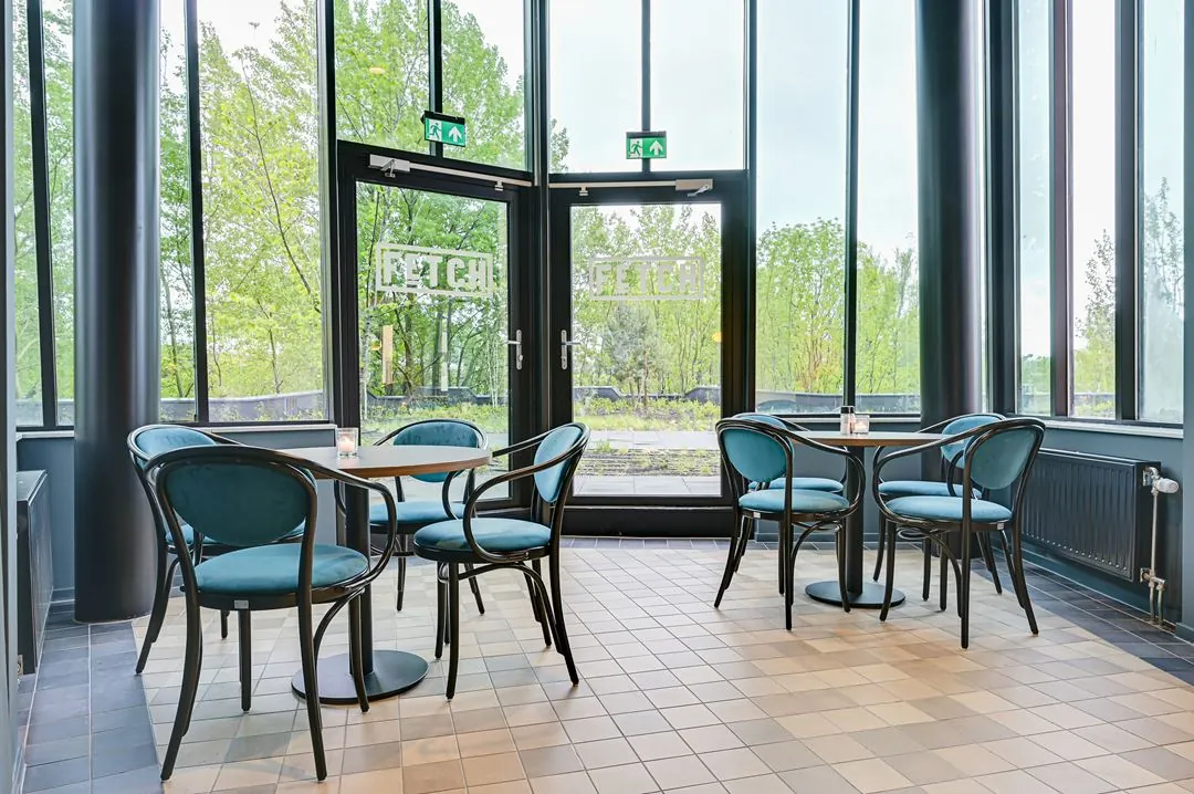Modern ingerichte zithoek met blauwe stoelen en ronde tafels in de lobby van Fetch op Kingsfordweg, met uitzicht op groene bomen door grote ramen.