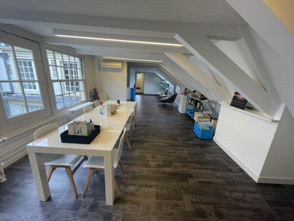 Moderne kantoorruimte aan de Herengracht met schuine plafonds, een lange witte tafel en grote ramen met uitzicht op klassieke gevels.