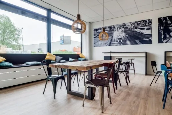 Moderne kantineruimte met houten tafel, industriële stoelen, hoge ramen en een zwart-wit stadsfoto aan de muur.