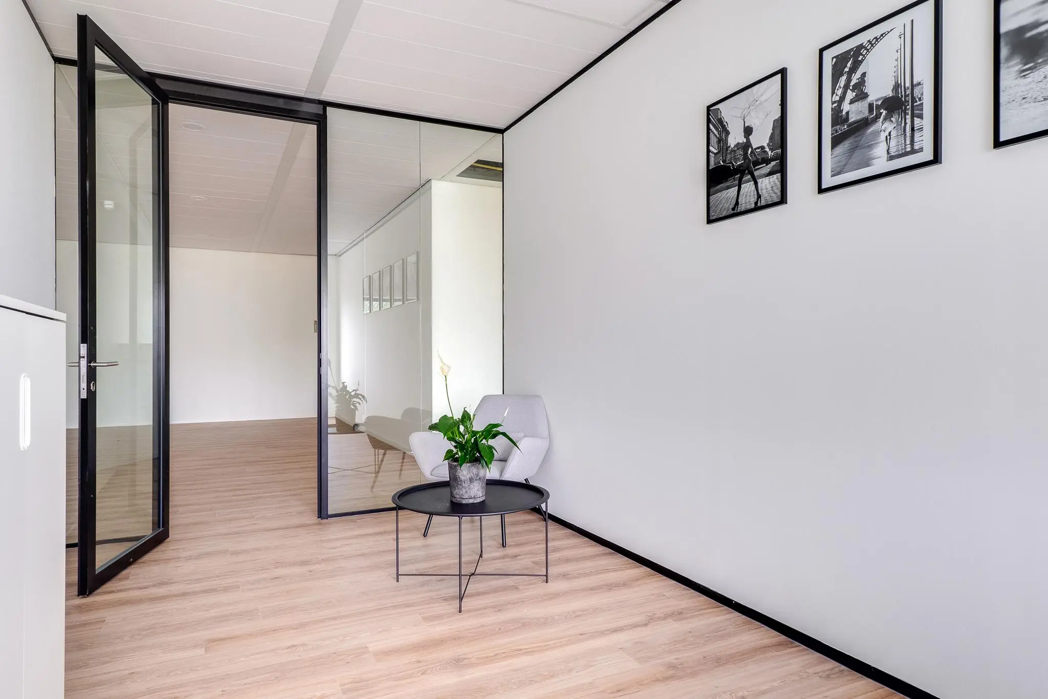 Moderne wachtruimte met een grijze fauteuil, een zwart bijzettafeltje met kamerplant en drie zwart-witfoto’s aan de muur.