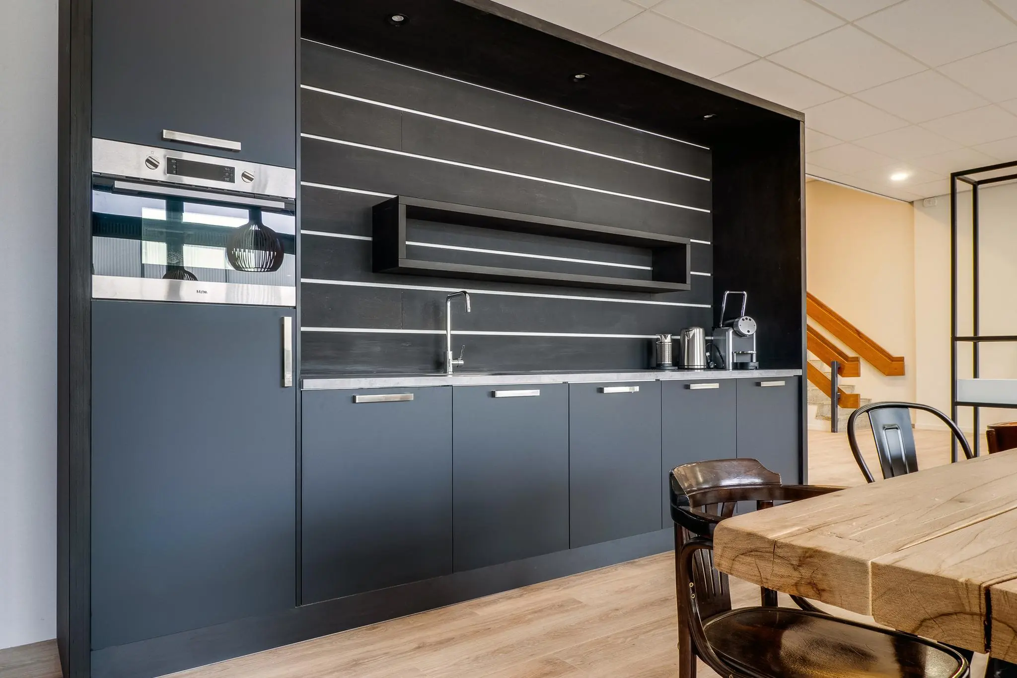 Moderne kantoorkeuken met donkergrijze kasten, een ingebouwde oven en een houten eettafel.
