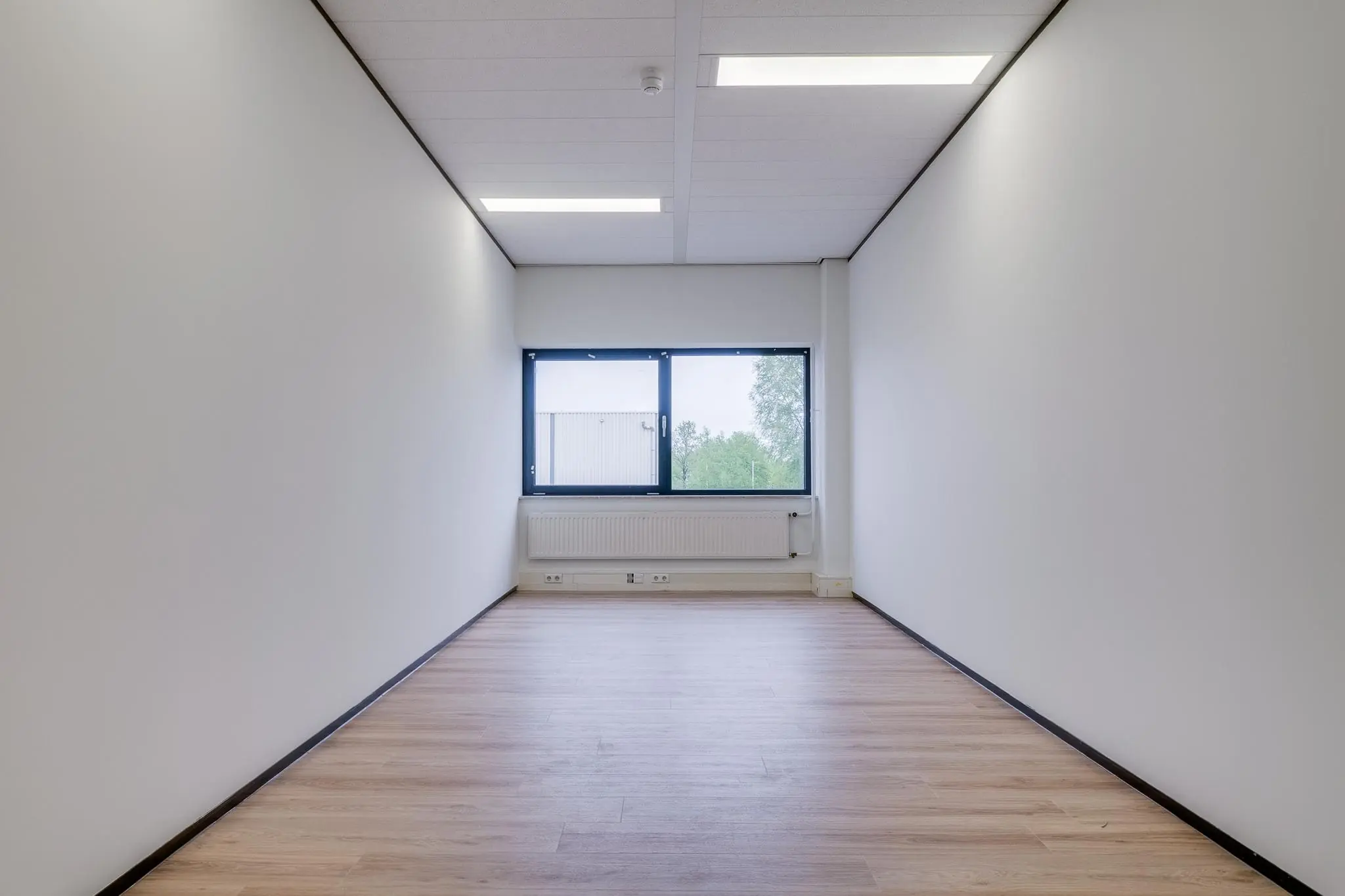 Lege, smalle kantoorruimte met witte muren, een houten vloer en een raam aan het einde.