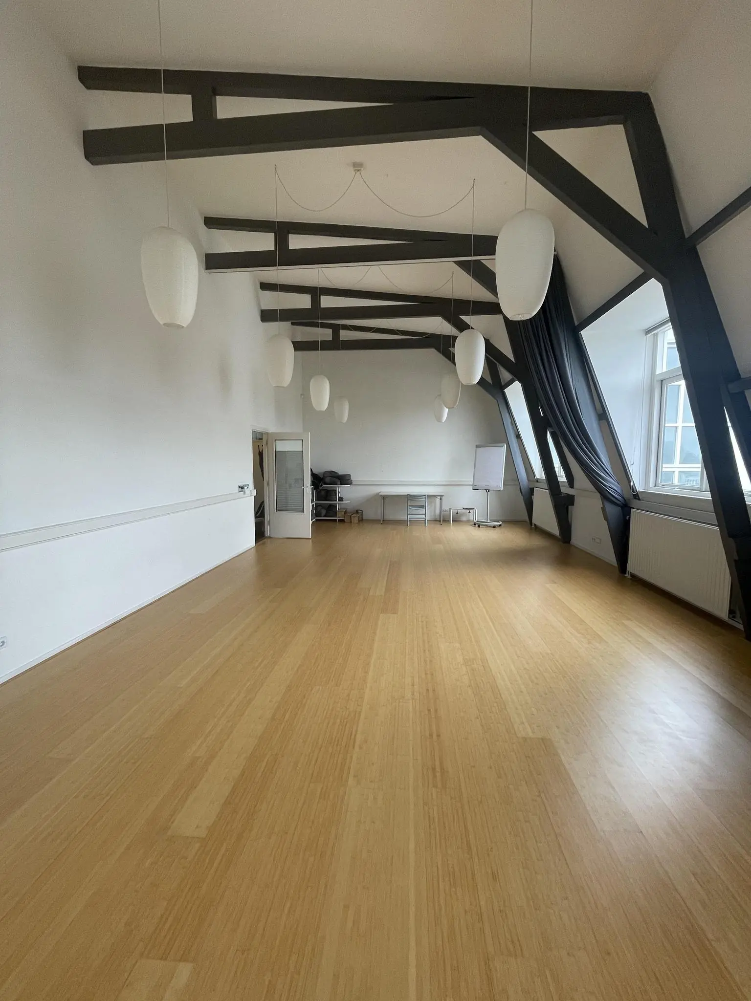 Lichte, ruime zaal aan de Raadhuisstraat met houten vloer, hoge balkenplafonds en hangende lampen.