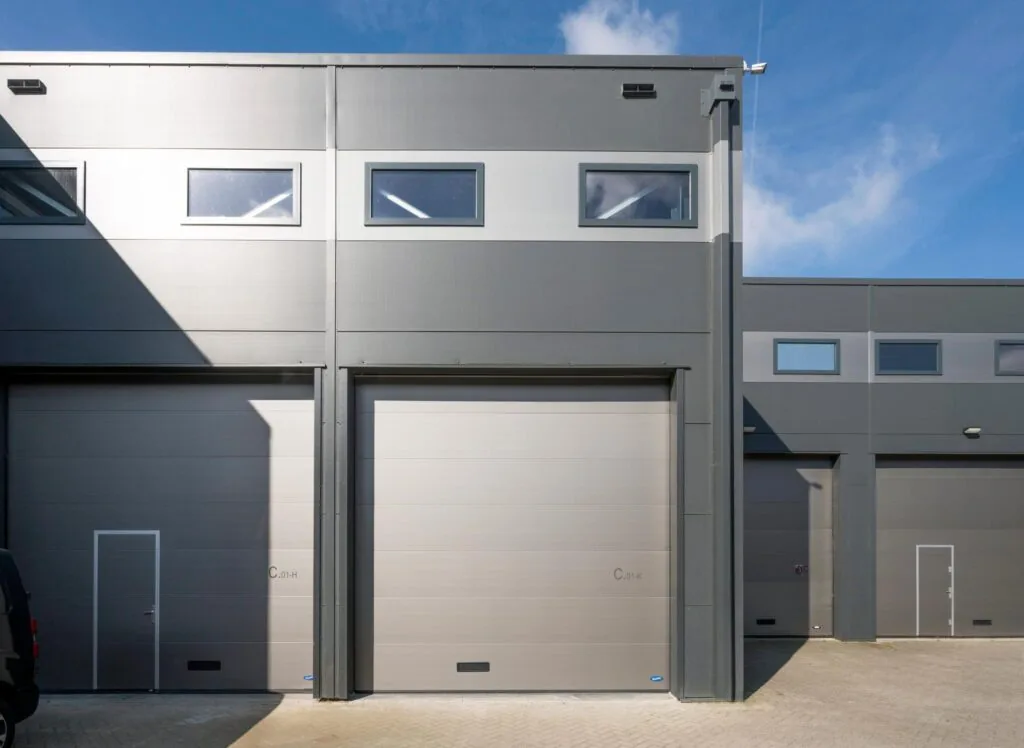 Grijze bedrijfsunits met hoge garagedeuren en bovenramen aan de Aalsmeerderweg, gefotografeerd op een zonnige dag.