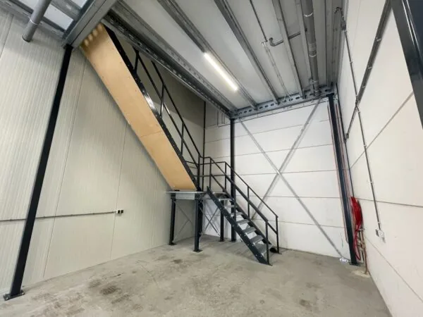 Industriële metalen trap in een loods aan de Aalsmeerderweg met zicht op betonnen vloer en leidingen aan het plafond.