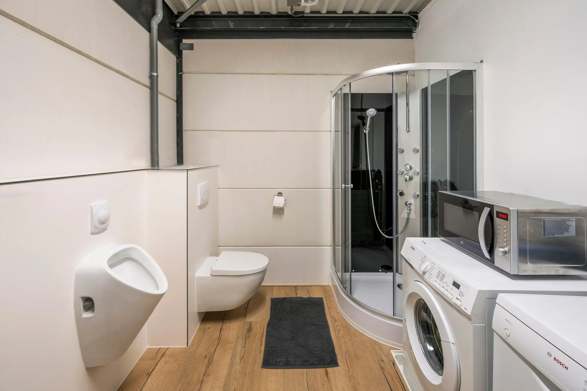 Moderne badkamer met douchecabine, toilet, urinoir, wasmachine, magnetron en houten vloer.