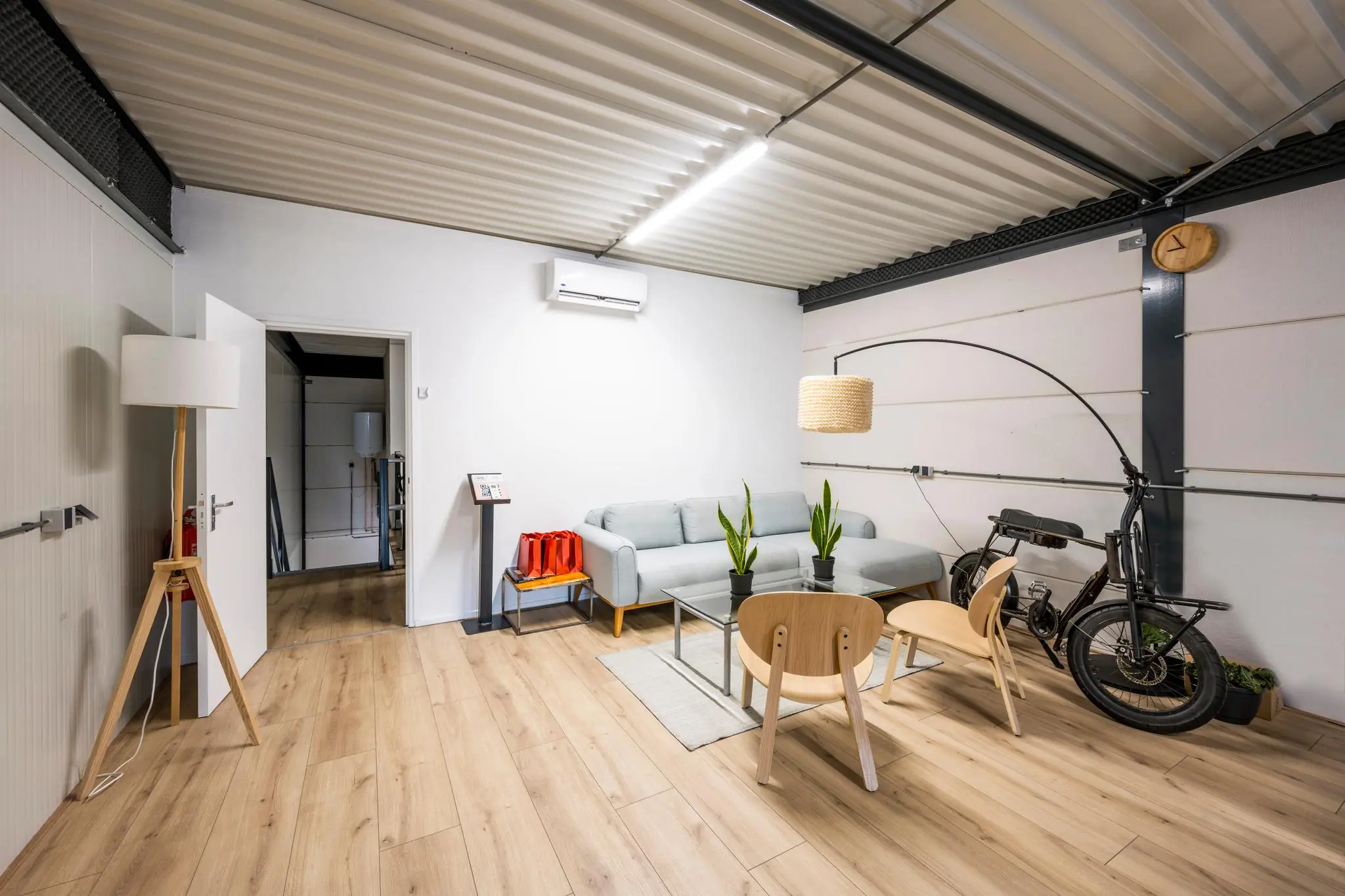 Moderne, strak ingerichte wachtruimte met een grijze bank, houten vloer, stoelen, planten en een elektrische fiets aan de muur op de Aalsmeerderweg.
