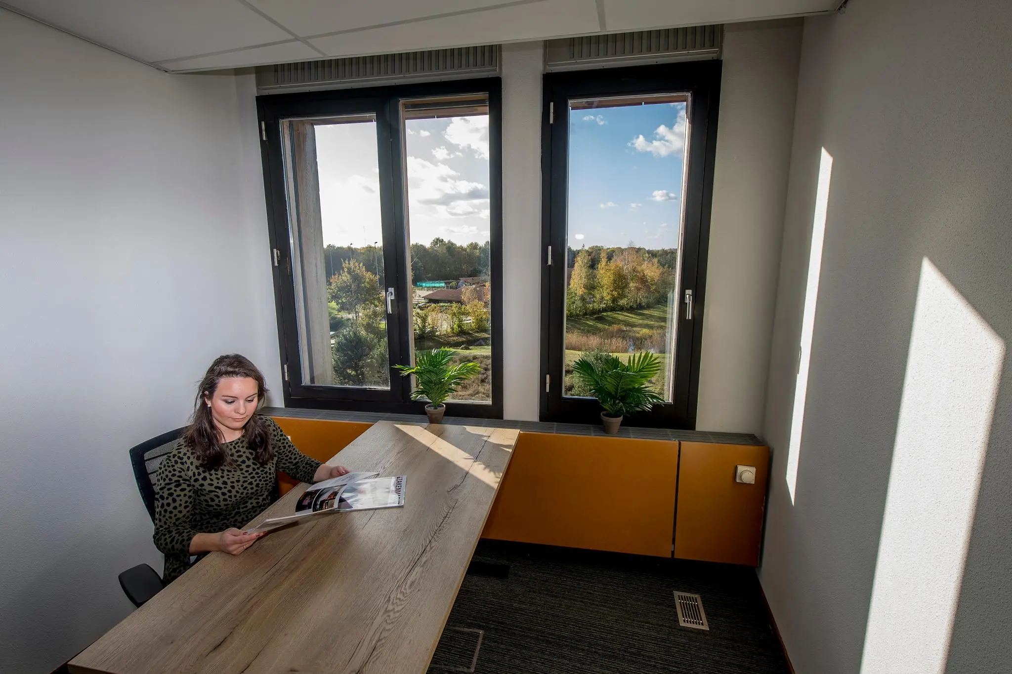 Vrouw leest een tijdschrift aan een bureau in een modern kantoor aan de Fokkerstraat met uitzicht op een groen landschap.
