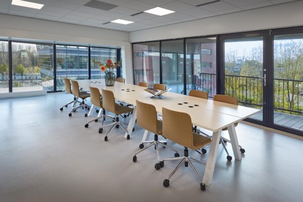 Vergaderruimte met lange tafel, moderne stoelen en uitzicht op kantoorgebouwen aan de Joop Geesinkweg.