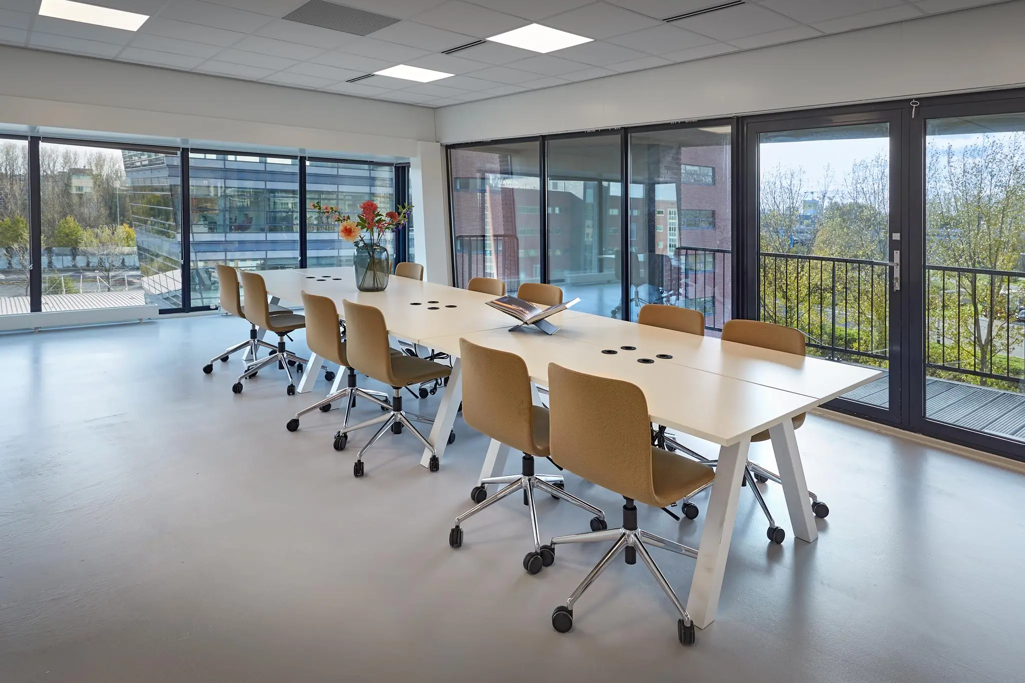Vergaderruimte met lange tafel, moderne stoelen en uitzicht op kantoorgebouwen aan de Joop Geesinkweg.