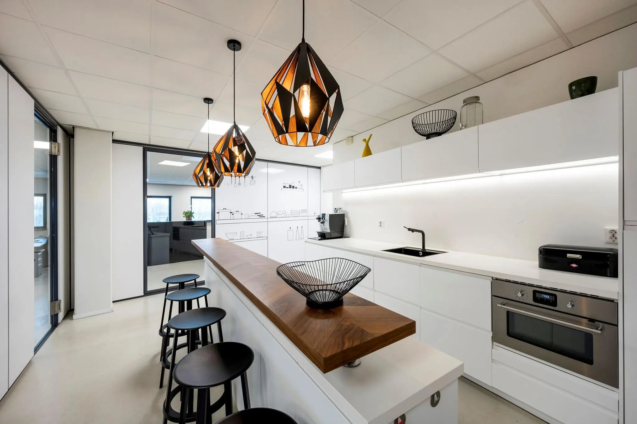 Moderne kantoorkeuken aan de Joop Geesinkweg met witte kasten, houten barblad, design verlichting en zwarte barkrukken.
