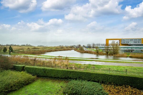 Uitzicht op een groen landschap met een waterpartij, een modern gebouw aan de Vareseweg en een deels bewolkte lucht.