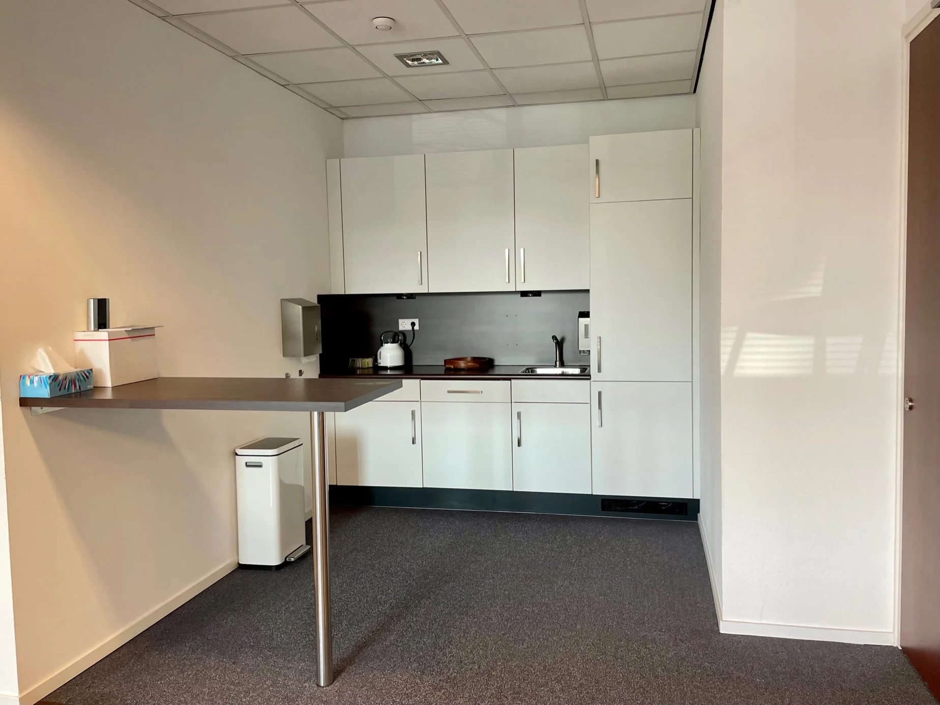 Kleine kantoorpantry met witte keukenkastjes, een aanrecht en een zwevende tafel tegen de muur.