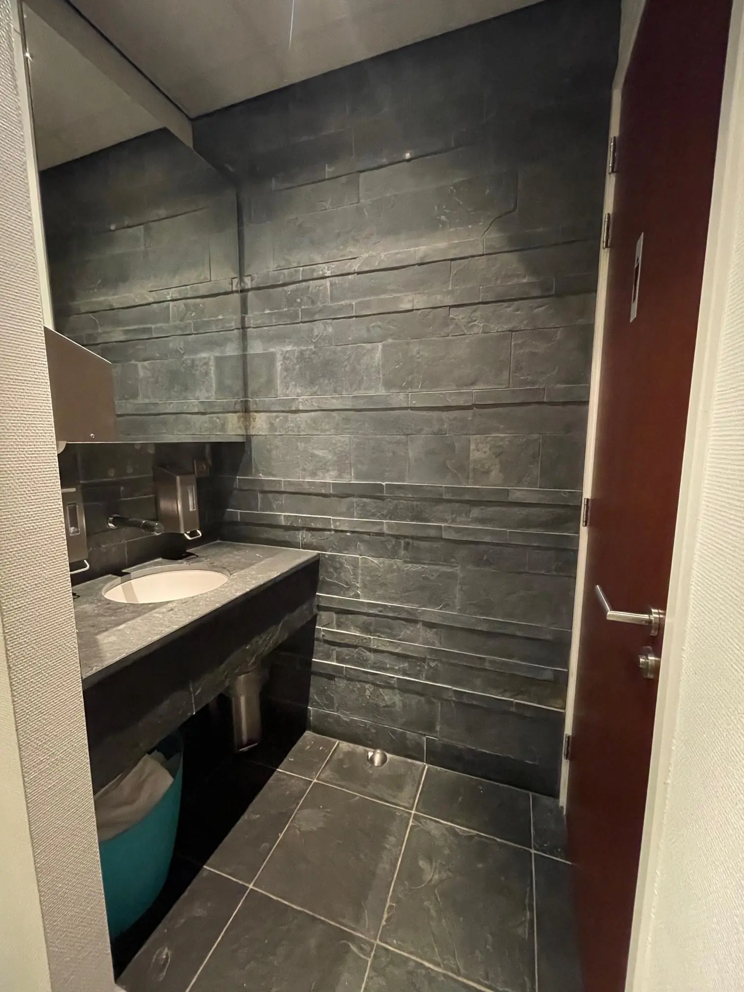 Moderne toiletruimte met donkere natuurstenen tegels, een wastafel met zeepdispensers en een gesloten deur.