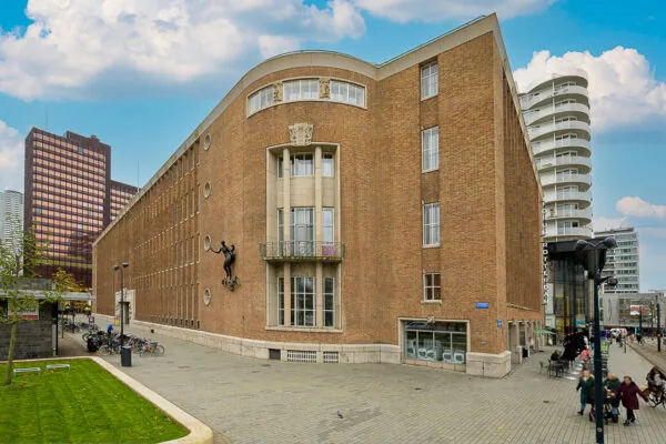 Bakstenen gebouw aan de Coolsingel in Rotterdam met een standbeeld aan de gevel en moderne hoogbouw op de achtergrond.