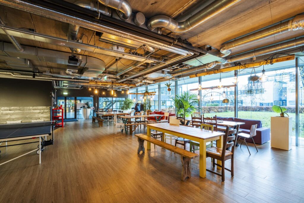 Moderne gemeenschappelijke ruimte aan de Naritaweg met houten tafels, zitgedeeltes, veel planten en industriële plafondbuizen.