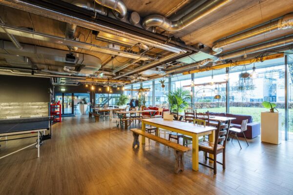 Moderne gemeenschappelijke ruimte aan de Naritaweg met houten tafels, zitgedeeltes, veel planten en industriële plafondbuizen.