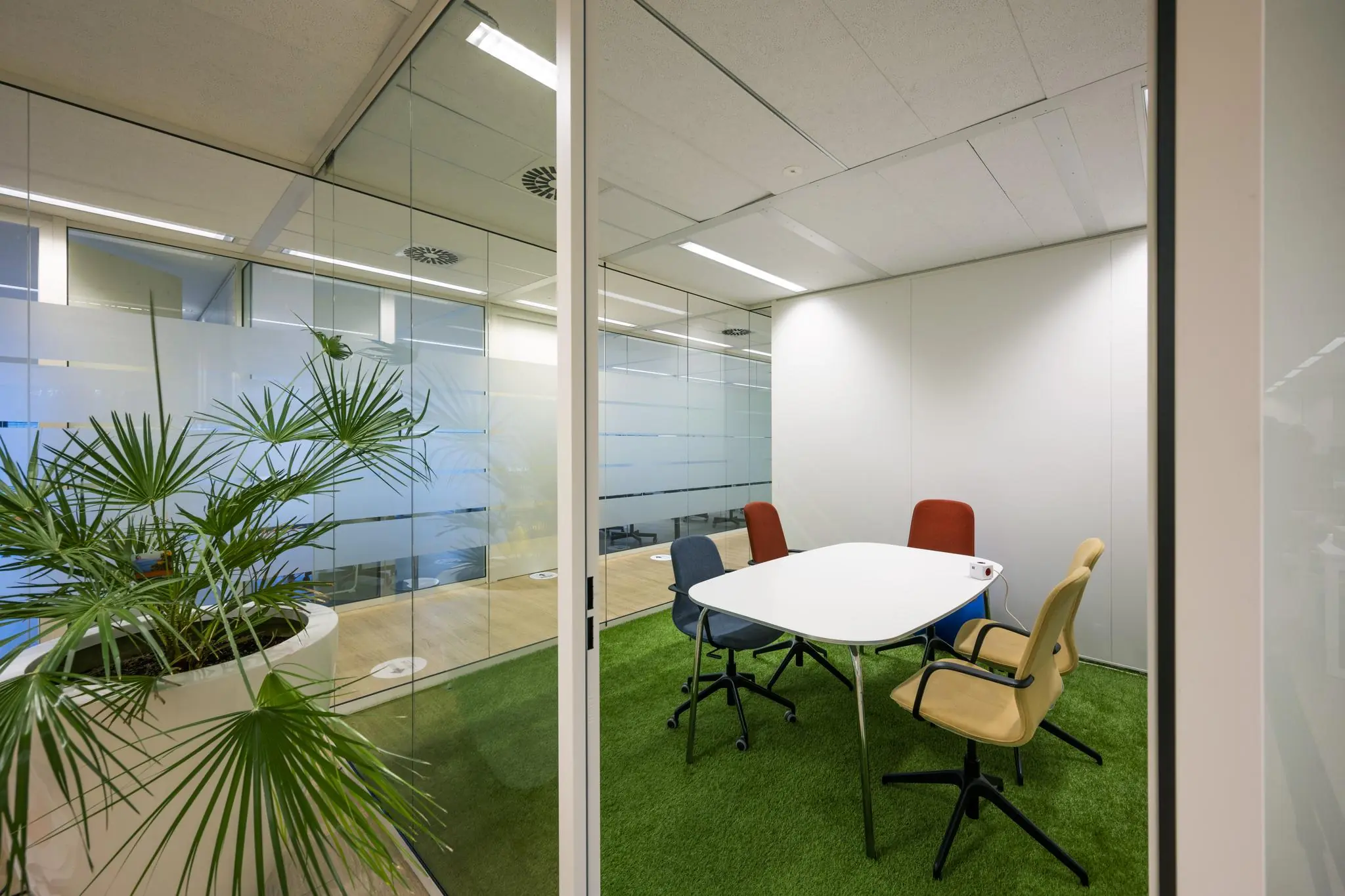 Kleine vergaderruimte aan de Naritaweg met glazen wanden, vier kleurrijke stoelen en kunstgrasvloer.