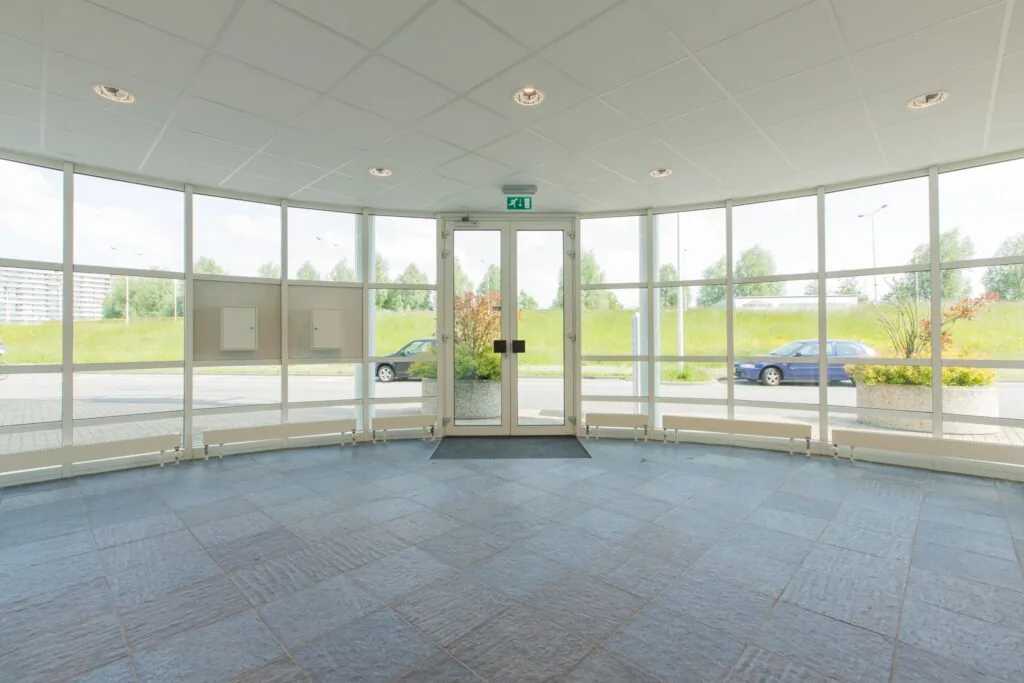 Ingang met glazen deuren en ramen van een kantoorpand aan het Rhijnspoor, met uitzicht op geparkeerde auto's en groenstrook.