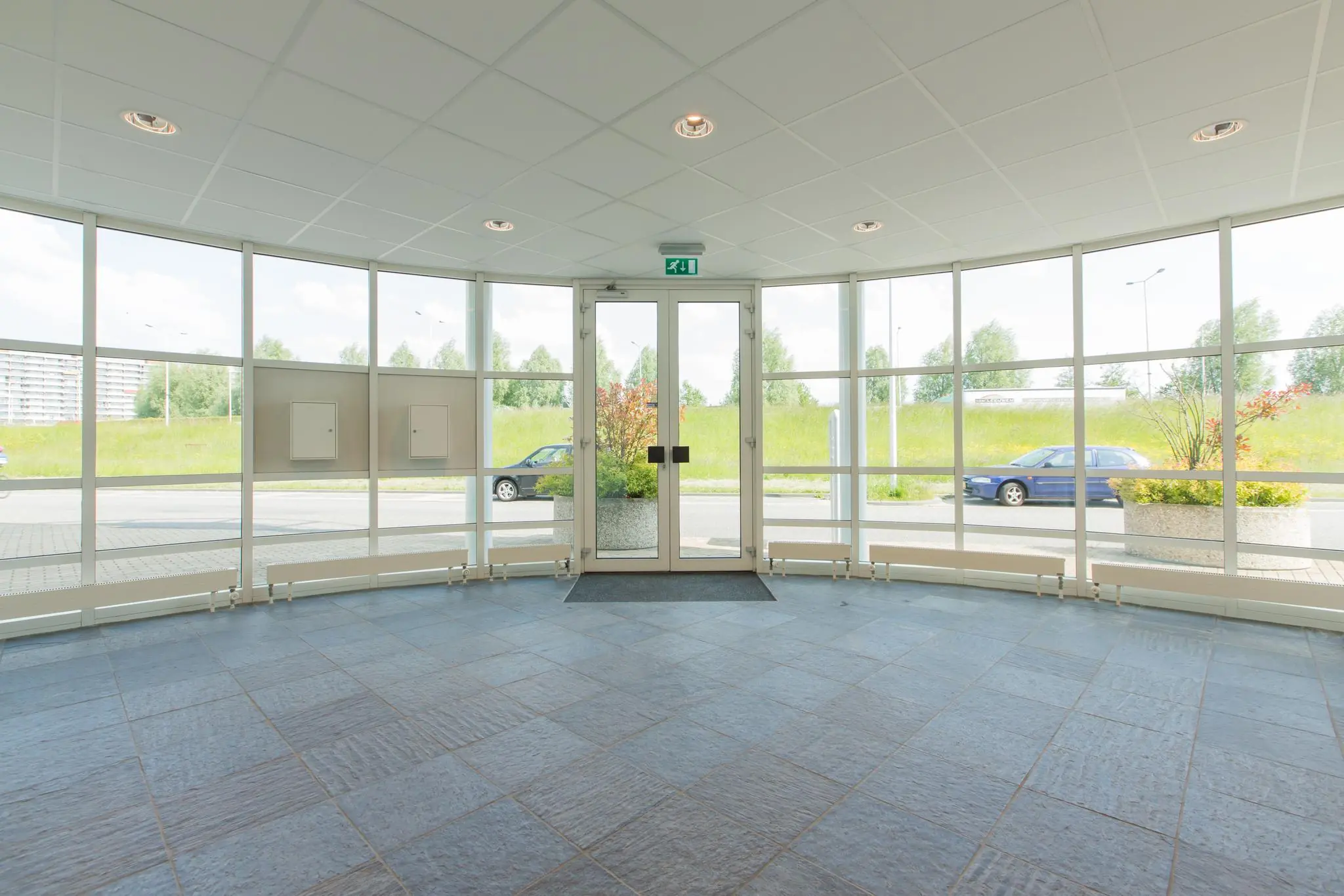 Ingang met glazen deuren en ramen van een kantoorpand aan het Rhijnspoor, met uitzicht op geparkeerde auto's en groenstrook.