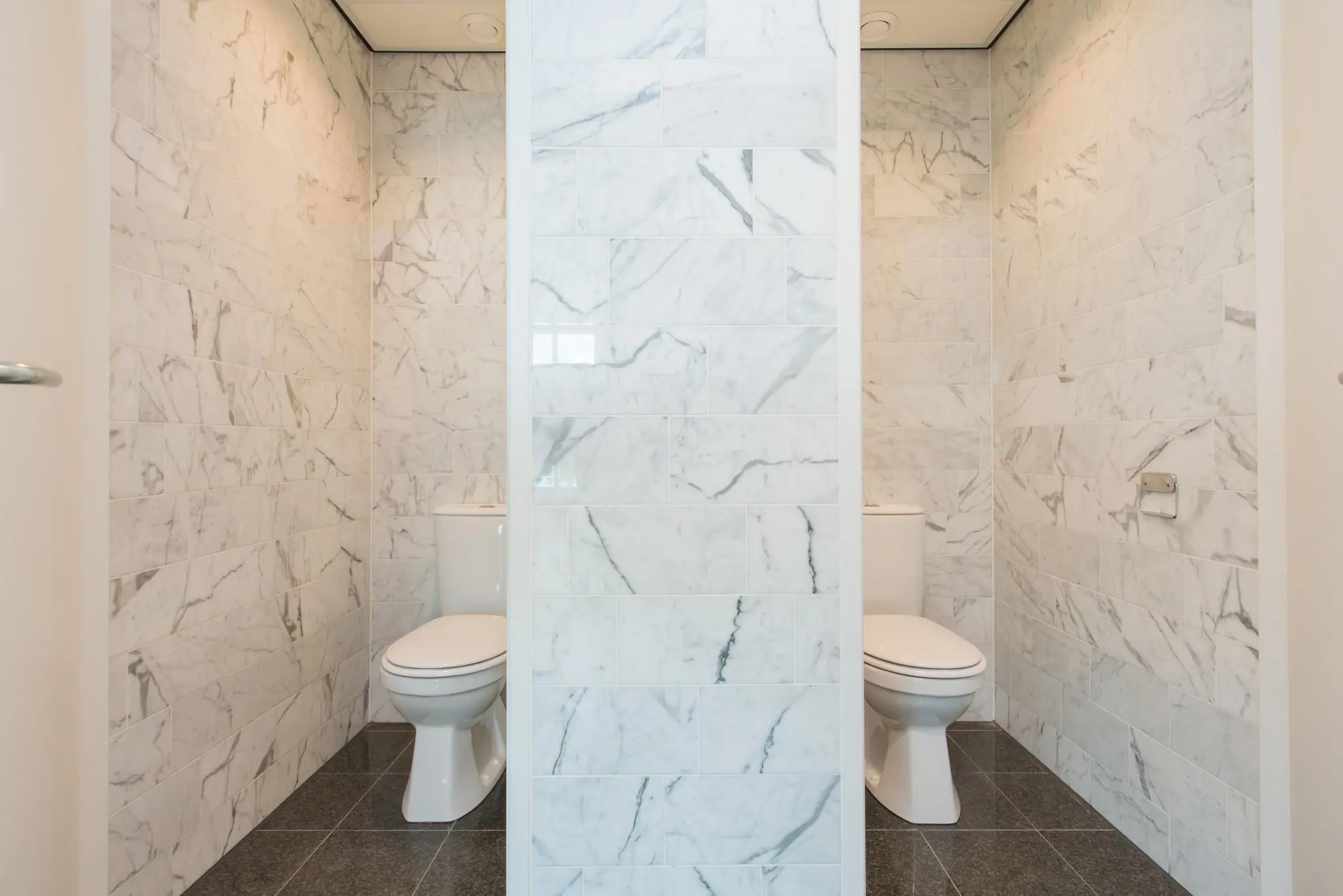 Twee moderne toiletruimtes met marmeren wanden, gescheiden door een tussenmuur.