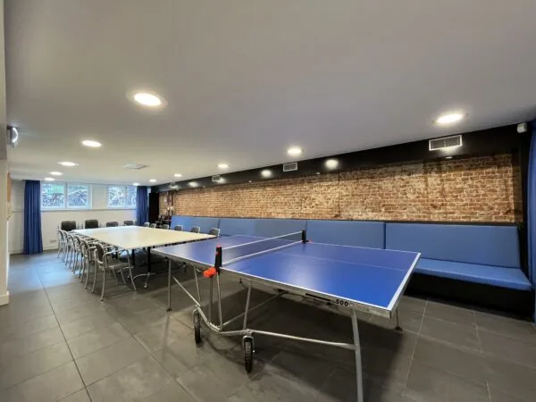 Vergaderruimte aan de Ramstraat met opklapbare tafels, een tafeltennistafel en een lange blauwe zitbank tegen een bakstenen muur.