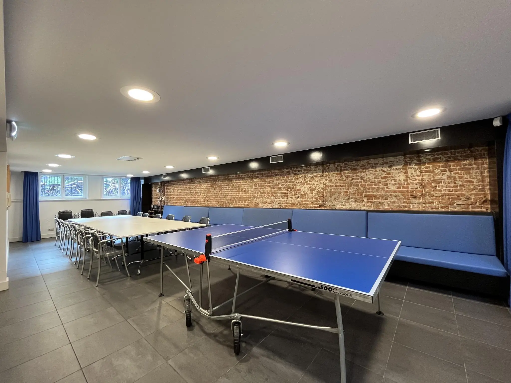Vergaderruimte aan de Ramstraat met opklapbare tafels, een tafeltennistafel en een lange blauwe zitbank tegen een bakstenen muur.