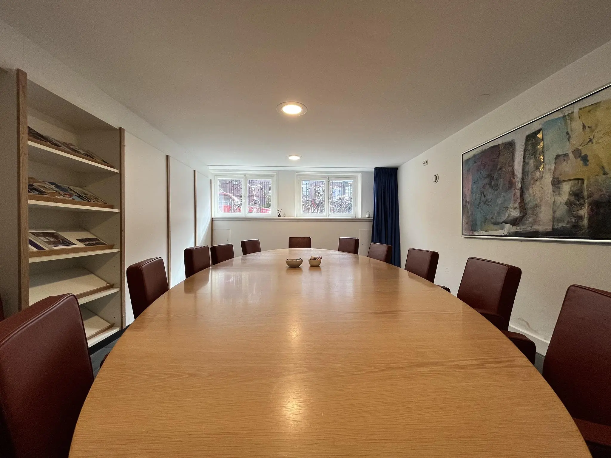 Vergaderruimte aan de Ramstraat met ovale houten tafel, rode stoelen, kunst aan de muur en een boekenkast.