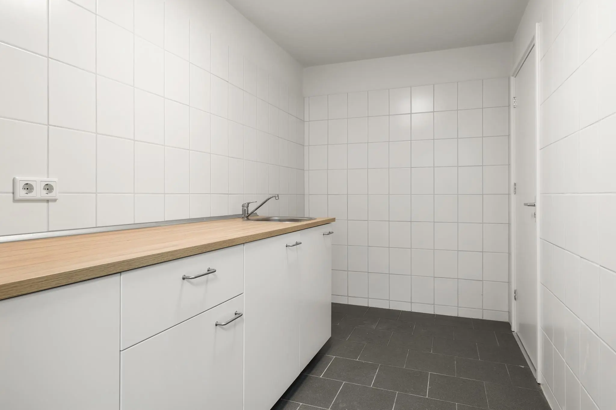 Strakke, wit betegelde bijkeuken met zwart tegelvloer, wit keukenblok en houten werkblad aan de Nijverheidsweg.