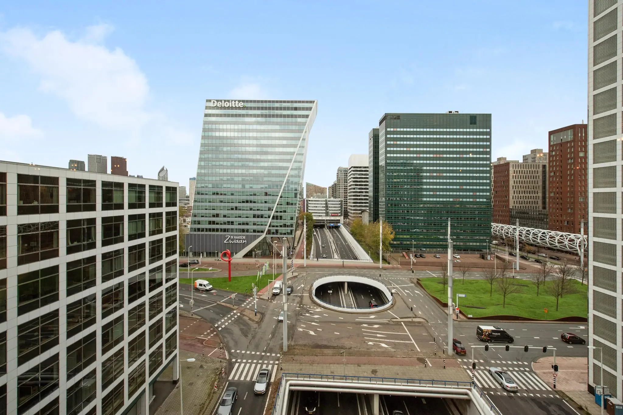 Stadszicht op de Schenkkade in Den Haag met moderne kantoorgebouwen, waaronder het Deloitte-gebouw "De Haagsche Zwaan", en een opvallende tunnelopening in het wegdek.