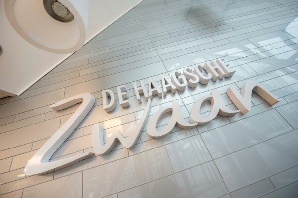 Wanddecoratie met de tekst "De Haagsche Zwaan" op een glanzende tegelmuur in een modern gebouw.