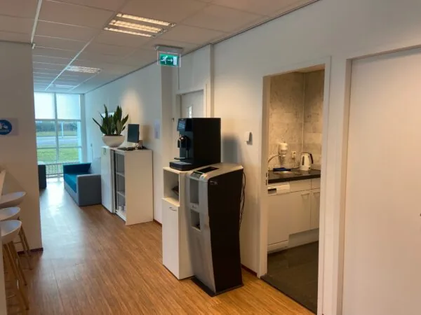 Kleine kantoorpantry met koffie- en waterautomaat, zithoek en open deur naar een kitchenette in gebouw Rhijnspoor.