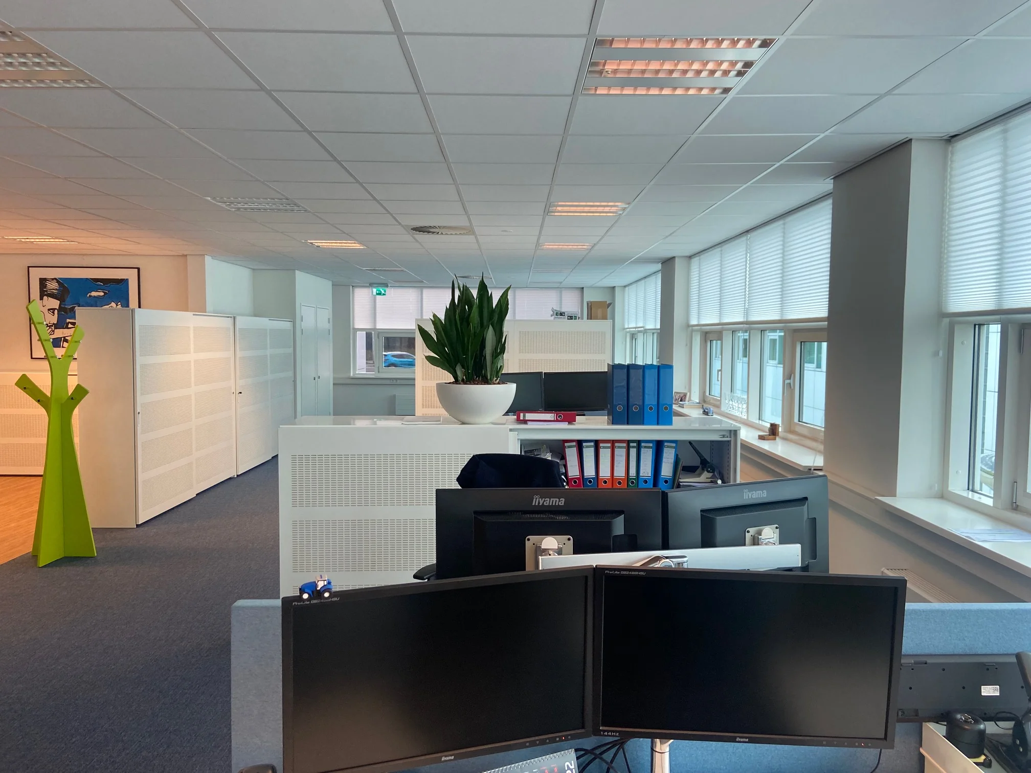 Moderne kantoorruimte aan het Rhijnspoor met bureaus, beeldschermen, archiefkasten en grote ramen met jaloezieën.