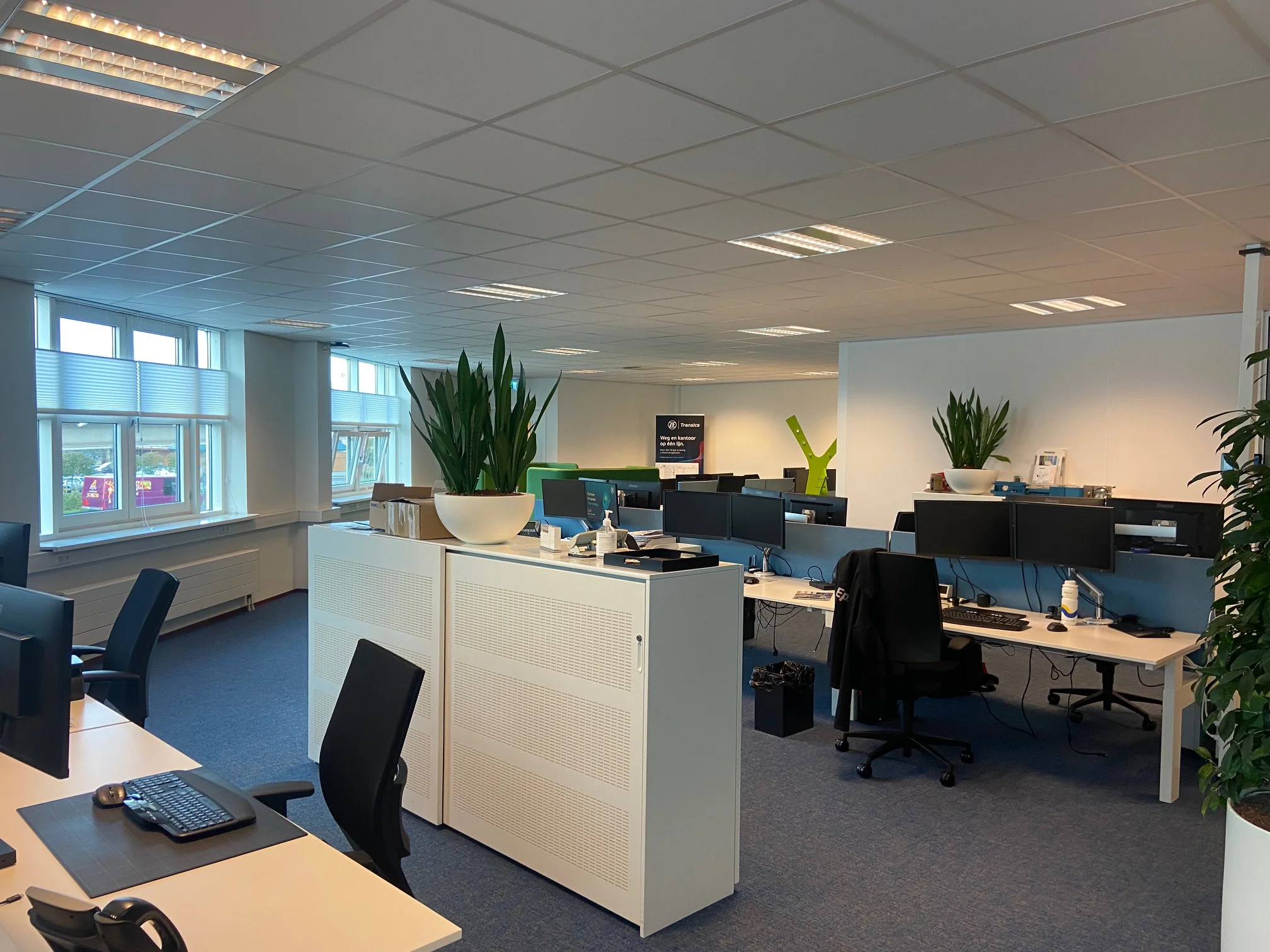 Modern kantoorpand aan het Rhijnspoor met meerdere werkplekken, bureaustoelen, beeldschermen en grote planten.