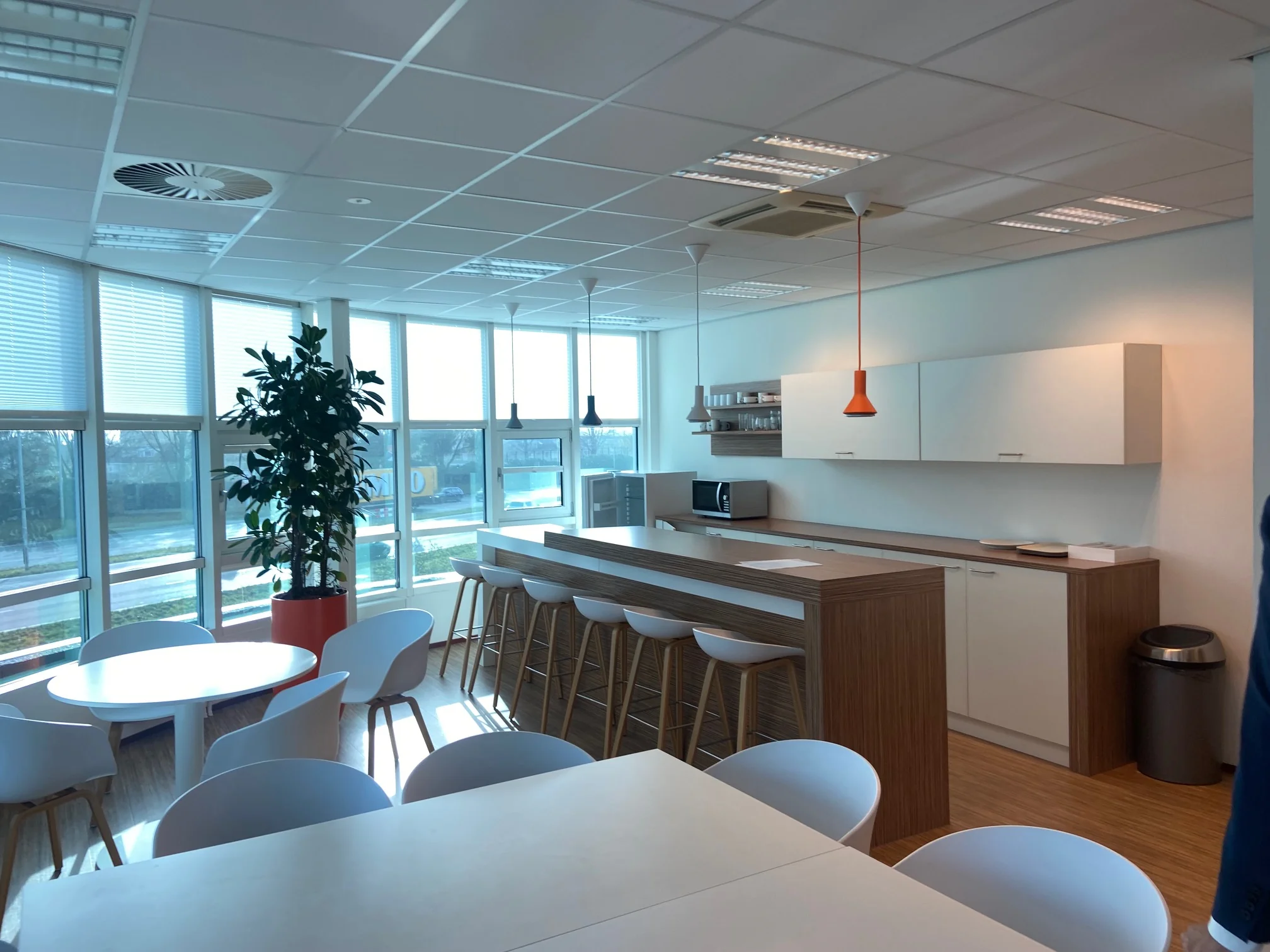 Moderne kantoorkeuken in het Rhijnspoor-gebouw met hoge ramen, een bar met krukken en een zithoek met ronde tafels.