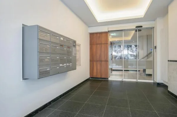 Ingang van een modern appartementen- of kantoorgebouw met grijze postvakken aan de muur en een schuifdeur van glas.