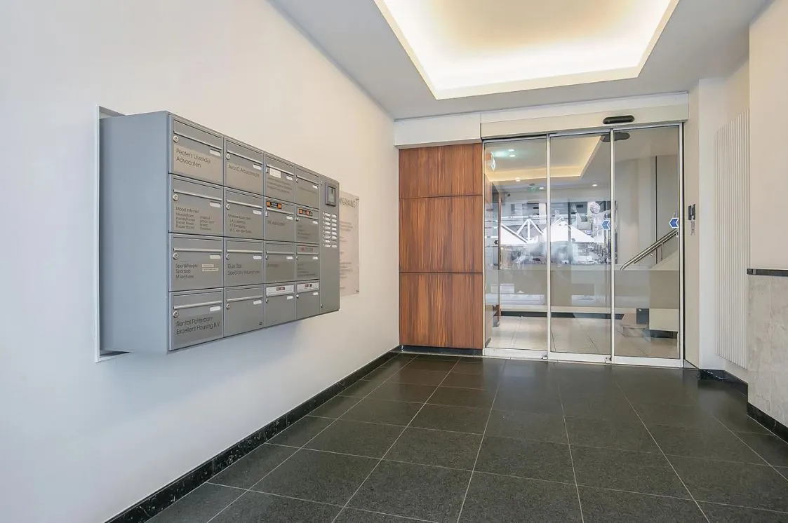 Ingang van een modern appartementen- of kantoorgebouw met grijze postvakken aan de muur en een schuifdeur van glas.