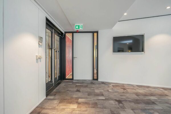 Moderne kantoorruimte met tapijtvloer, een glazen deur, een witte binnendeur met nooduitgangsbord en een flatscreen aan de muur.