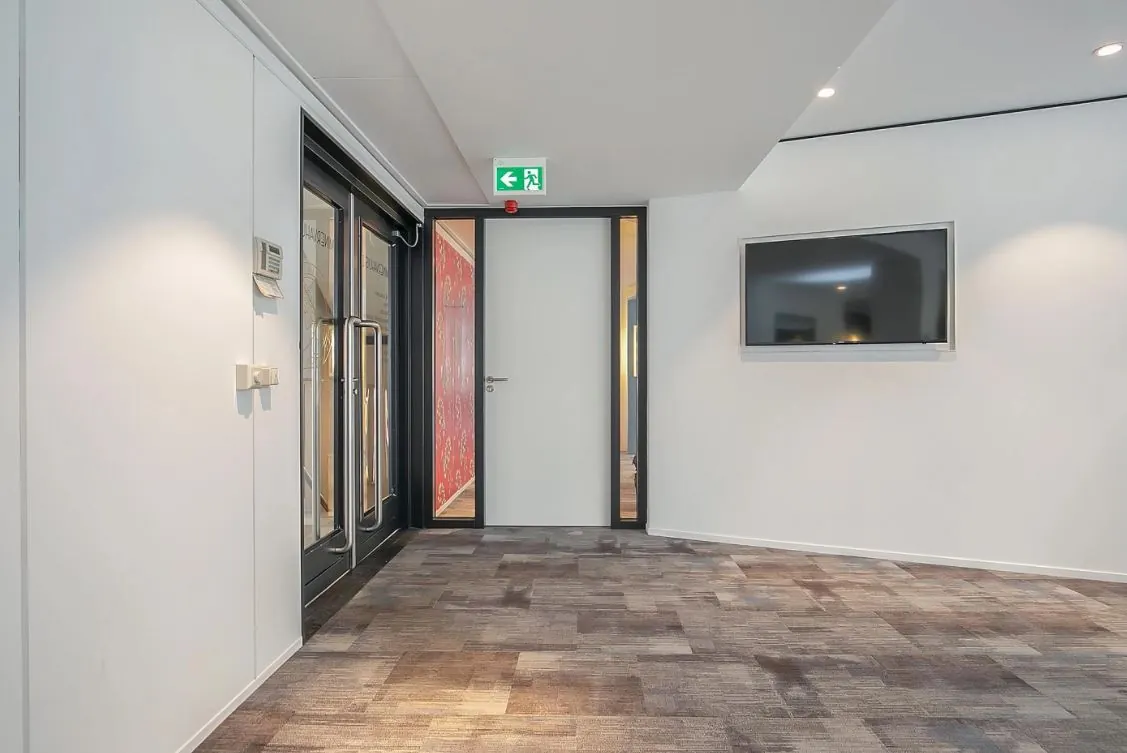 Moderne kantoorruimte met tapijtvloer, een glazen deur, een witte binnendeur met nooduitgangsbord en een flatscreen aan de muur.