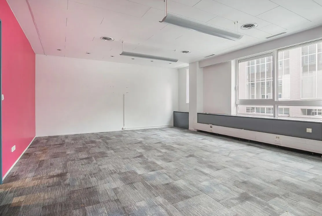 Leeg kantoorpand met grijze tapijttegels, een felroze muur en grote ramen aan de rechterzijde.