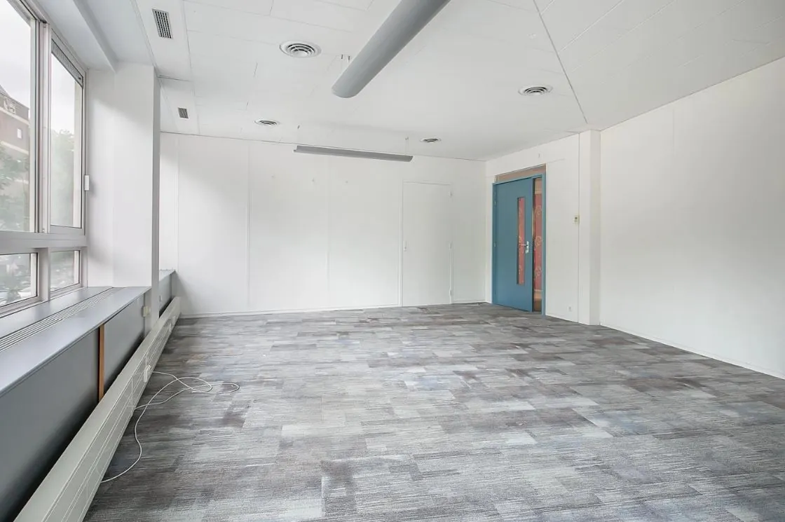 Lege kantoorruimte met grote ramen, grijs tapijt en een blauwe deur aan de achterzijde.