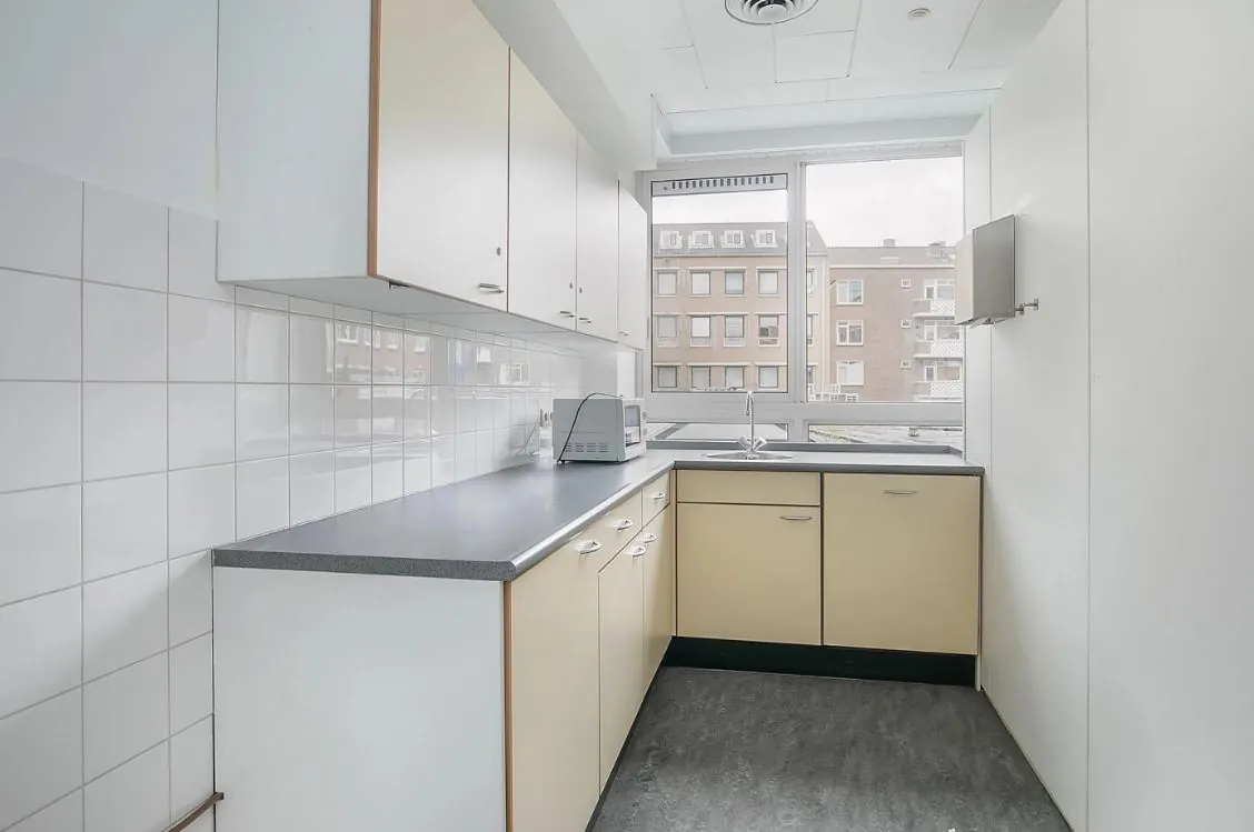 Lichte hoekkeuken met beige kastdeuren, witte wandtegels en een groot raam met uitzicht op een woonwijk aan de Meent.
