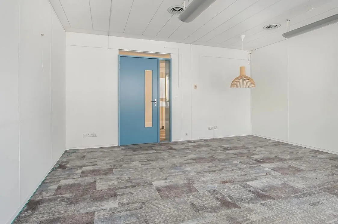 Lege kantoorruimte met grijs tapijt, witte muren, een blauwe deur en een houten hanglamp aan het plafond.