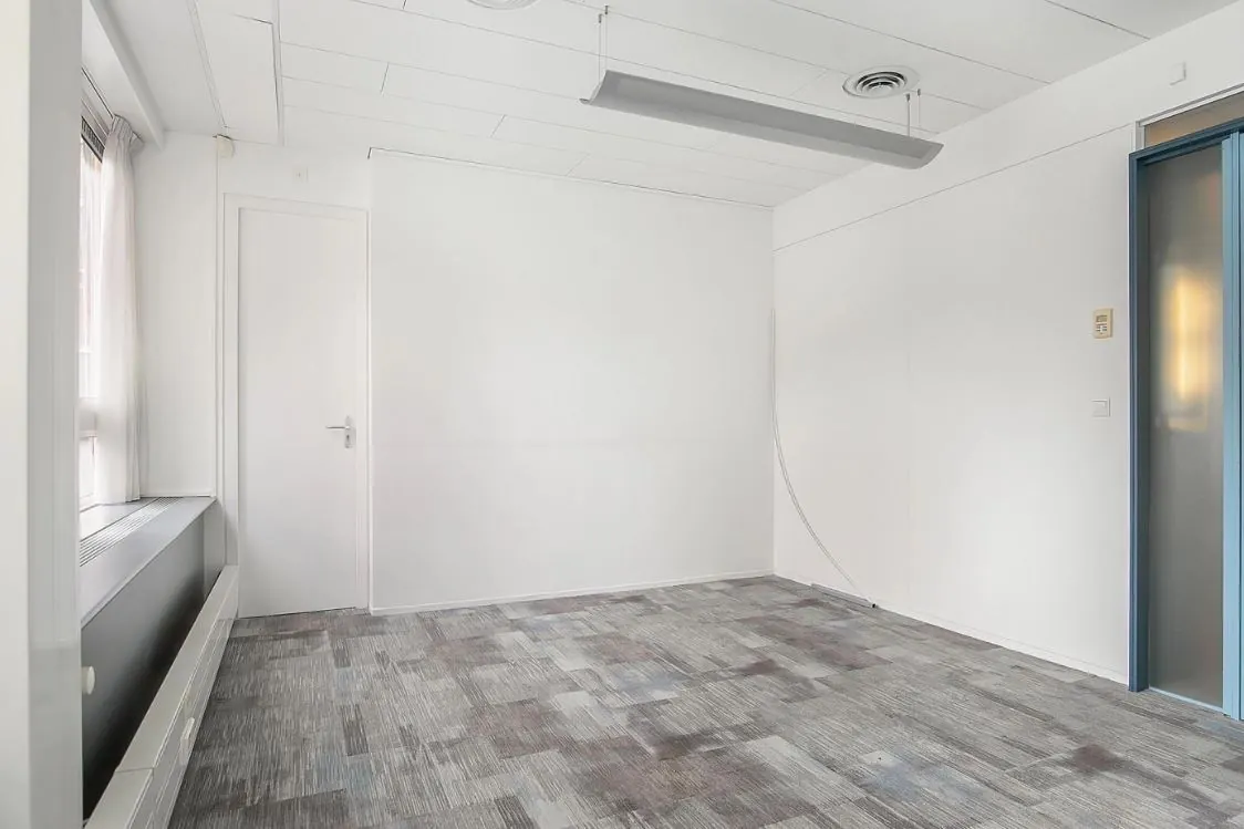 Lege witte kantoorruimte met grijs tapijt, een deur, raam en systeemplafondverlichting.